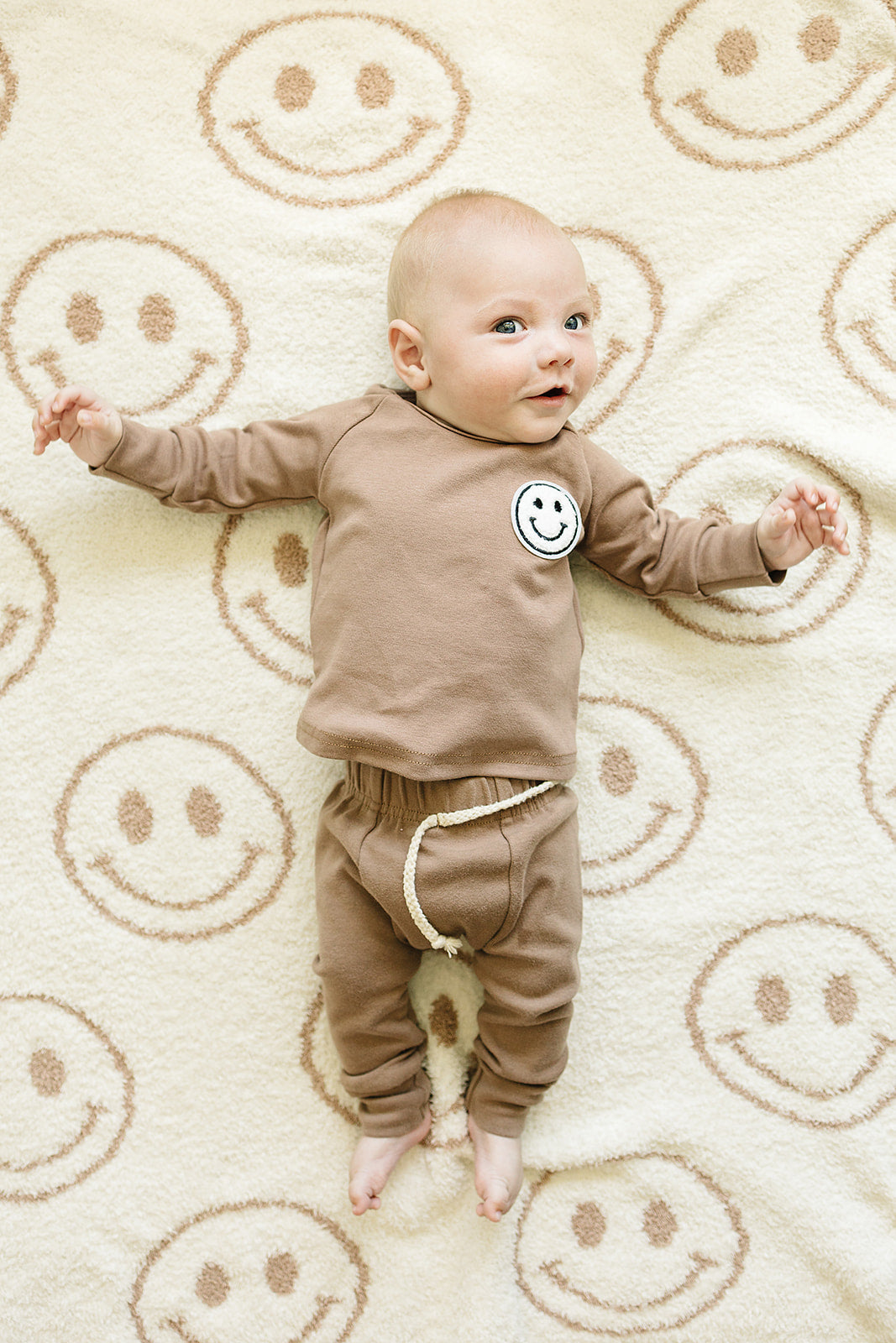 baby wearing brown two piece outfit lying on smiley face patterned plush blanket
