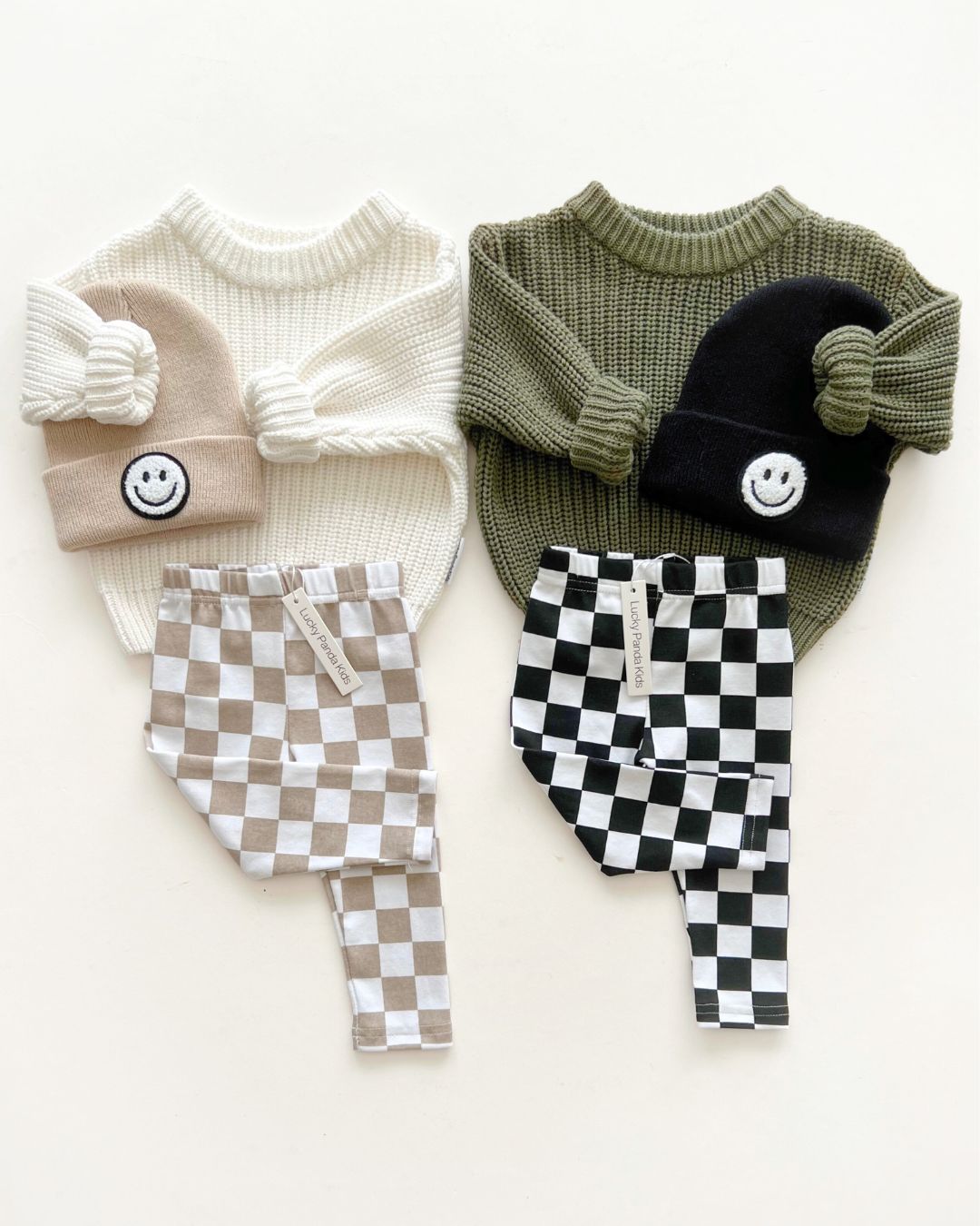 flat lay of two baby outfits with knit sweaters, smiley beanies, and checkered pants in neutral colors