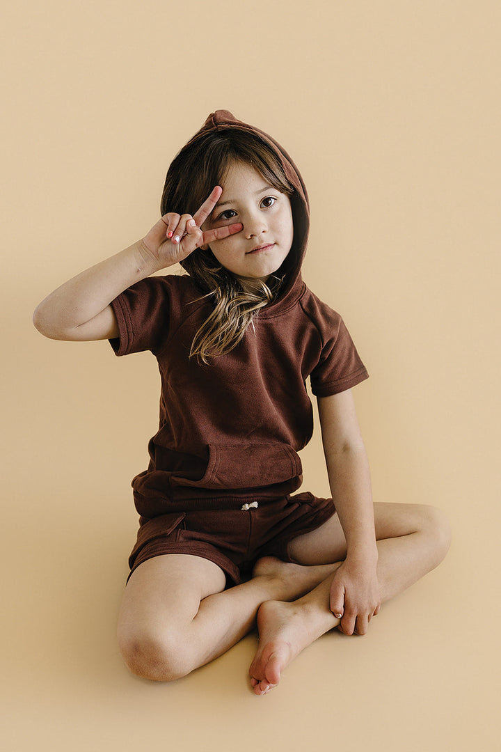 Child wearing a brown hoodie and shorts sitting on a beige background