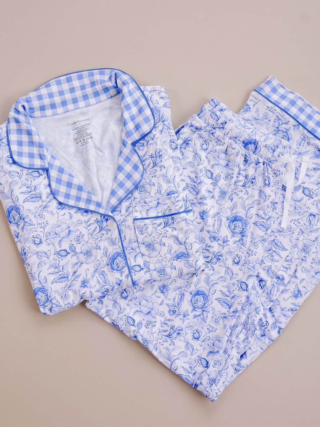 A two-piece pajama set laid flat on a beige background, featuring a blue and white gingham collar with blue piping and a matching blue floral print on a white base.