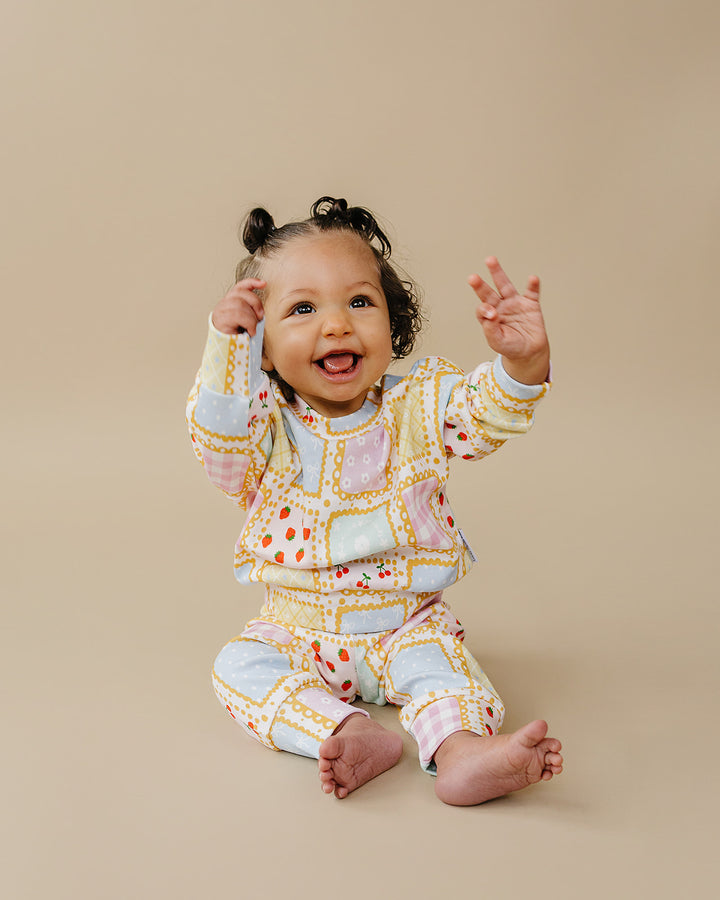 Smiling baby sitting with both hands raised in the Sweet Patchwork top and matching jogger pants.