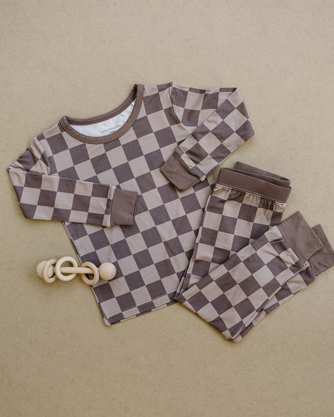 A baby pajama set consisting of a long-sleeve top and matching pants in a brown and beige checkerboard pattern, laid flat on a textured beige background next to a wooden teething ring toy.