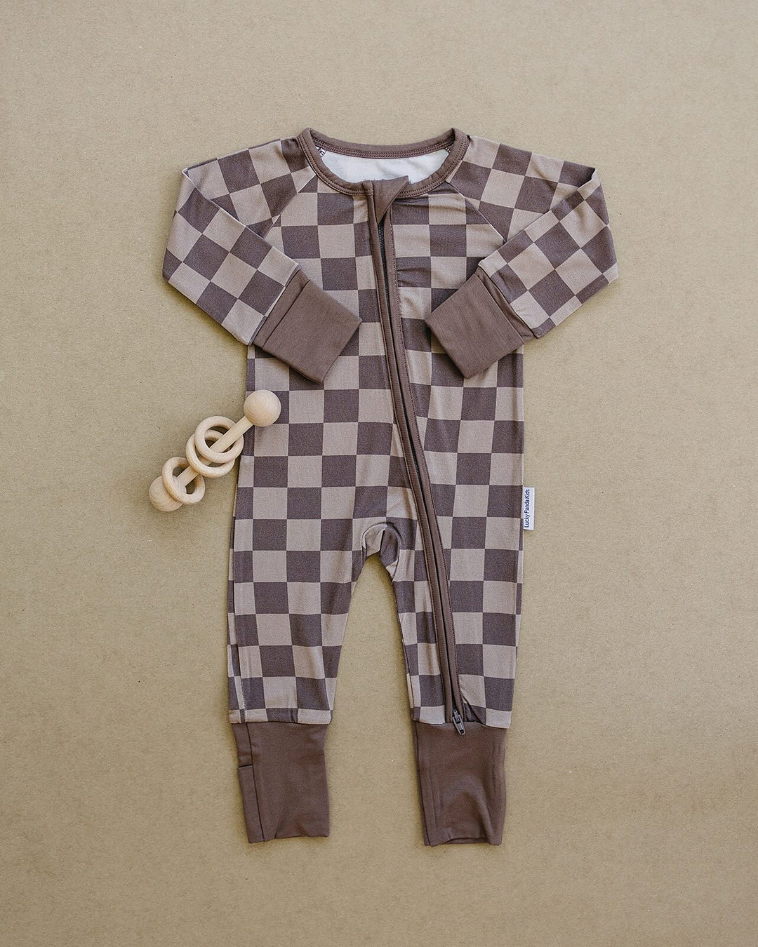 A baby long-sleeve zip-up footless sleeper with a brown and beige checkerboard pattern, laid flat on a textured beige background next to a wooden ring teether.