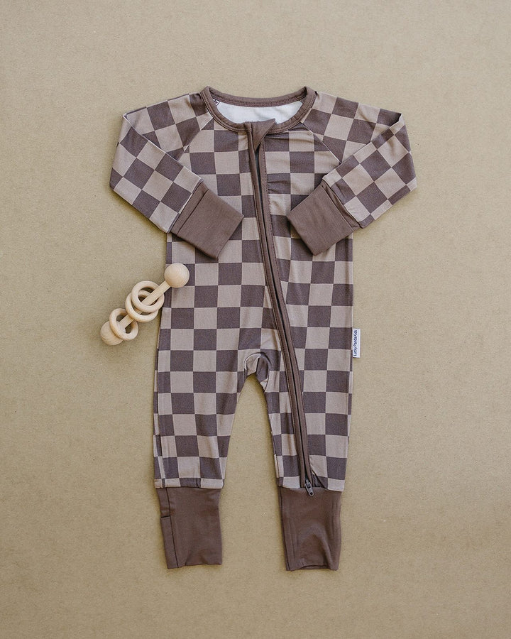 A baby long-sleeve zip-up footless sleeper with a brown and beige checkerboard pattern, laid flat on a textured beige background next to a wooden ring teether.