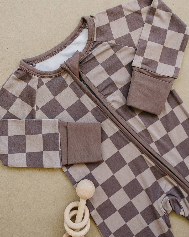 A baby footed sleeper with a brown and beige checkerboard pattern, laid flat on a neutral beige background with a wooden teething ring toy.