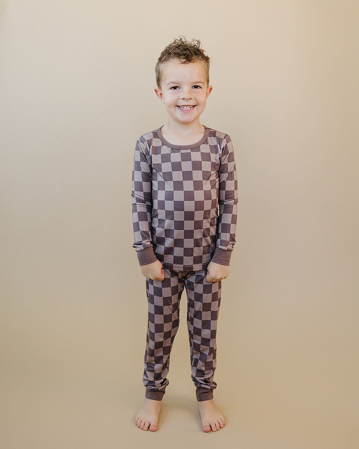 A young boy stands barefoot against a solid beige background, wearing a long-sleeve and matching pants pajama set with a brown and gray checkerboard pattern.