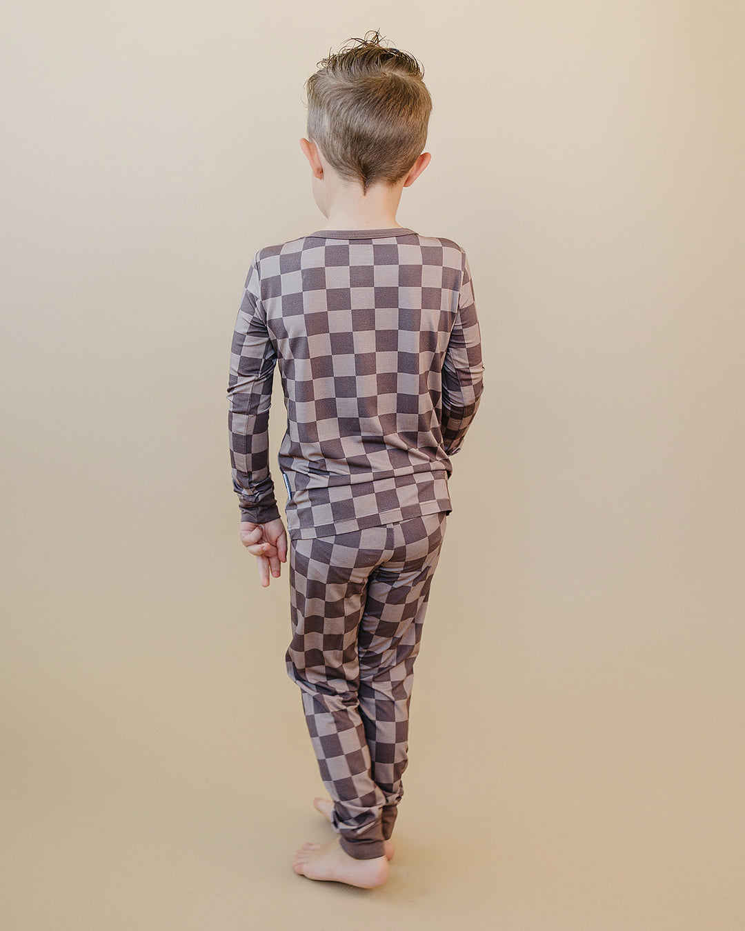 A child viewed from behind wearing a long-sleeve pajama set with a brown and gray checkerboard pattern, standing against a solid beige background.