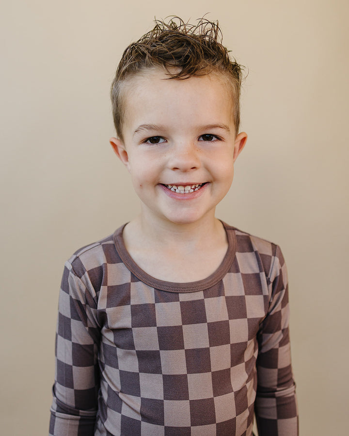 A young boy smiling at the camera, wearing a brown and beige checkered long-sleeve shirt against a solid beige background.