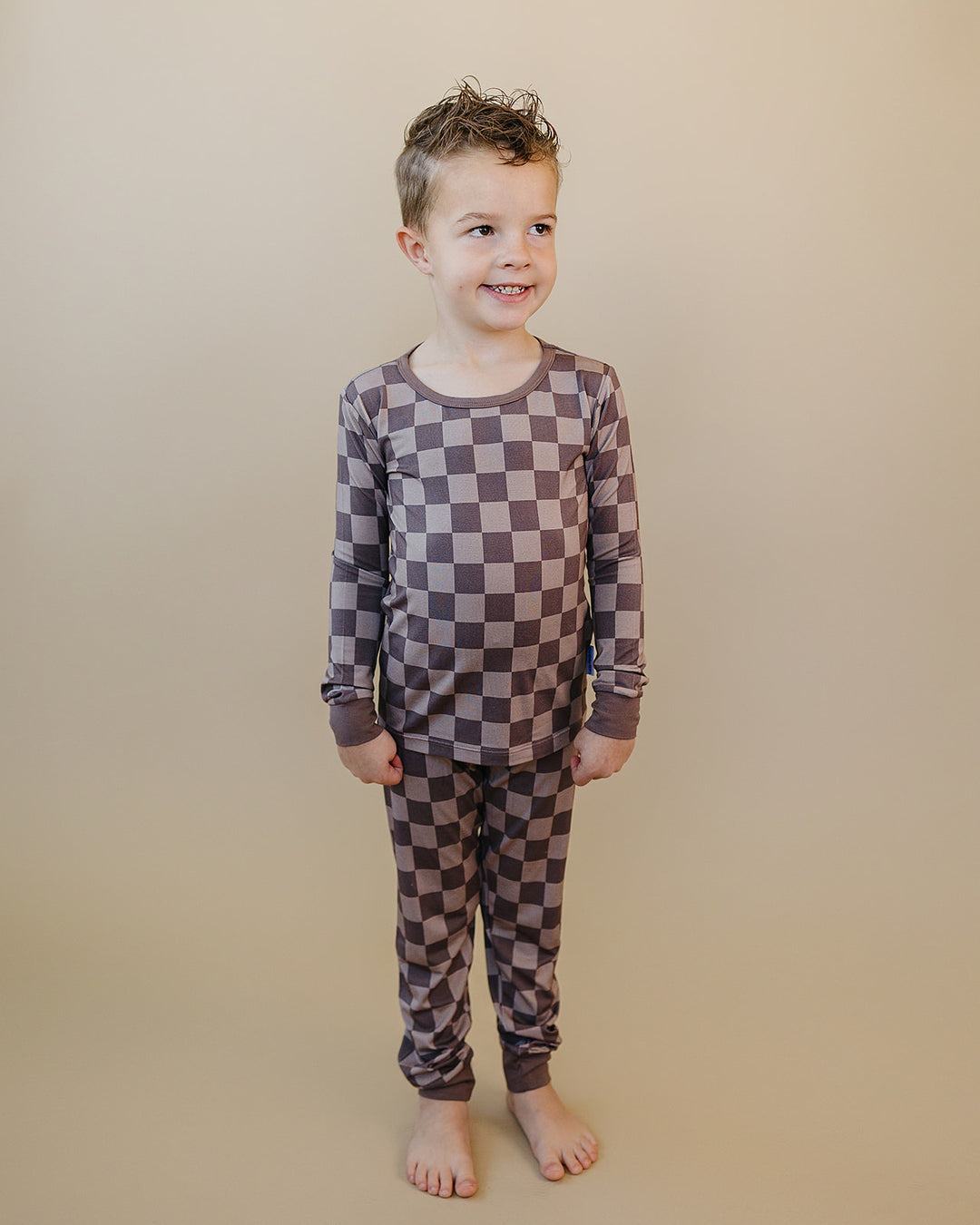 A young boy stands barefoot against a plain beige studio background, wearing a long-sleeve pajama set with a brown and gray checkerboard pattern.