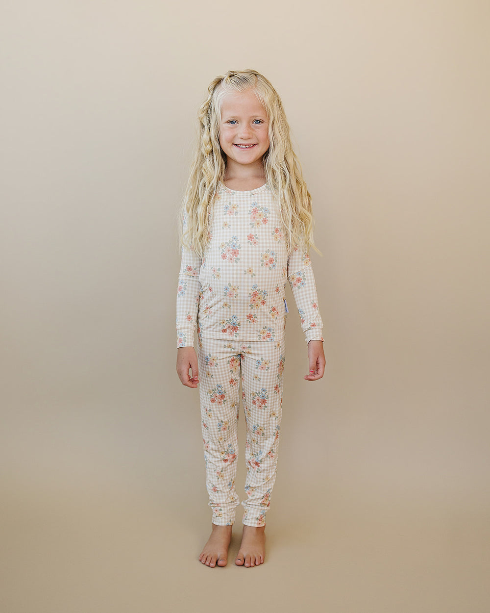 A young girl with long blonde hair stands barefoot against a solid beige studio background, wearing a long-sleeve matching pajama set with a small white gingham grid pattern and a scattered floral print in pink, blue, and yellow.