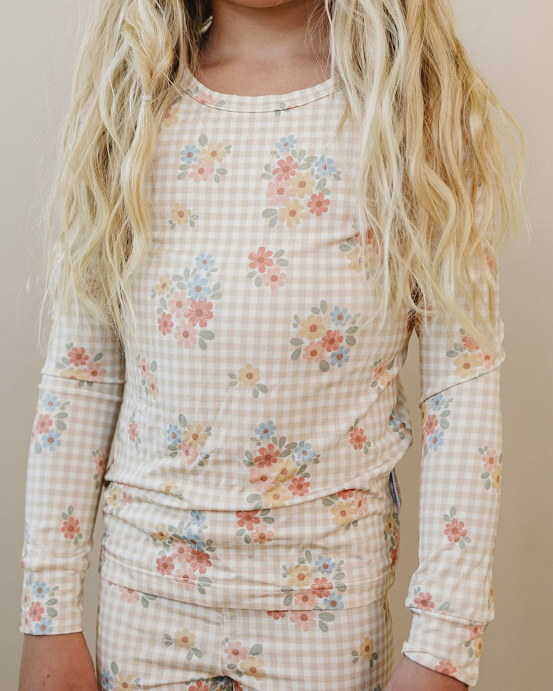 A close-up of a person wearing a long-sleeve pajama top with a beige gingham check pattern and a scattered floral print in pink, blue, yellow, and green. The image is cropped from the neck down, showing the model's blonde hair and the fit of the pajama top against a plain beige background.