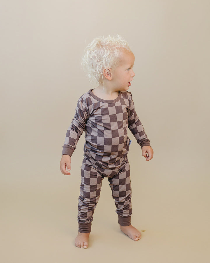 A toddler with curly blonde hair stands barefoot against a plain beige studio background, wearing a long-sleeve one-piece pajama set with a brown and taupe checkerboard pattern.