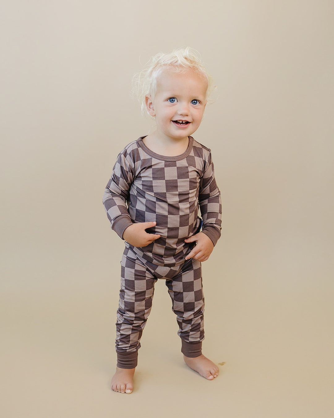 A smiling toddler with blonde hair stands barefoot against a solid beige studio background, wearing a long-sleeve and matching pants set in a brown and taupe checkerboard pattern.