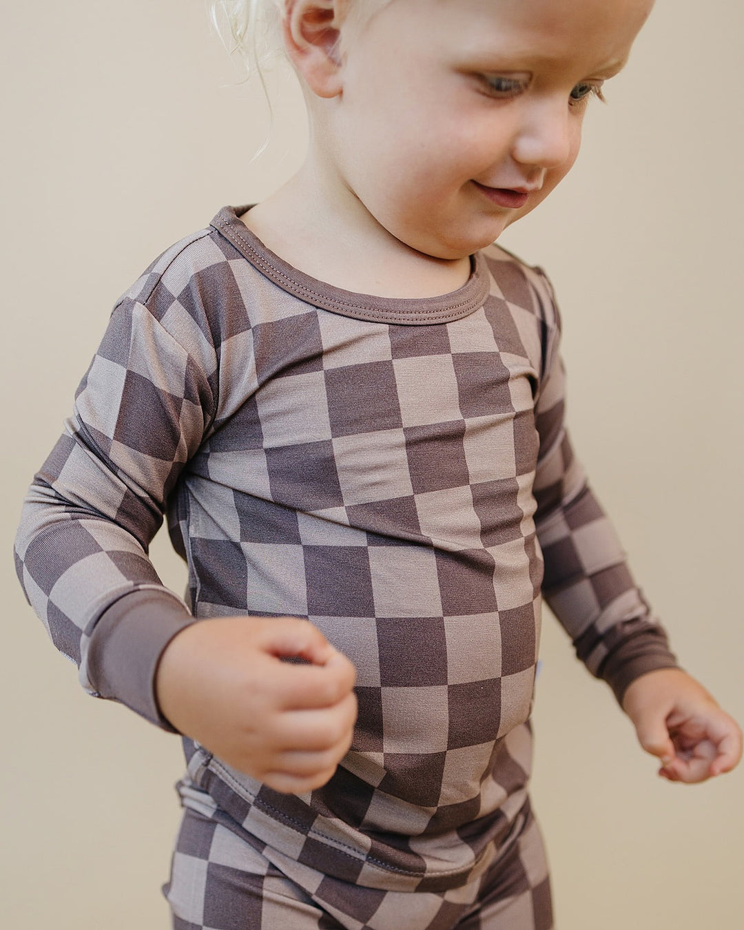 A toddler wearing a long-sleeve checkered pajama top in muted brown and gray tones, photographed against a plain beige background.