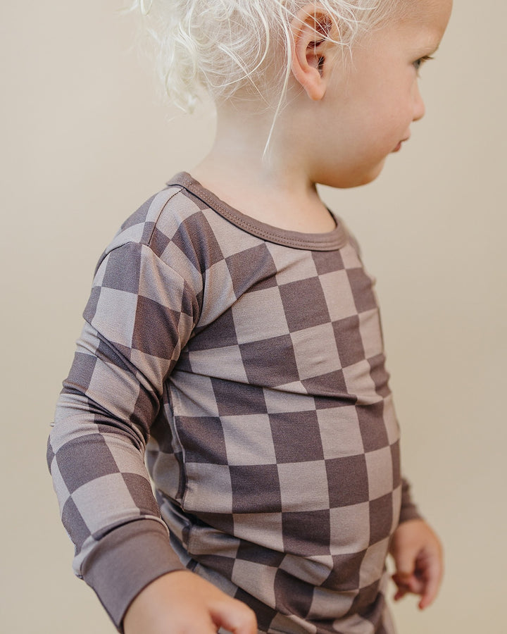 A toddler wearing a long-sleeve checkered shirt in muted brown and gray tones, shown in profile against a plain beige background.