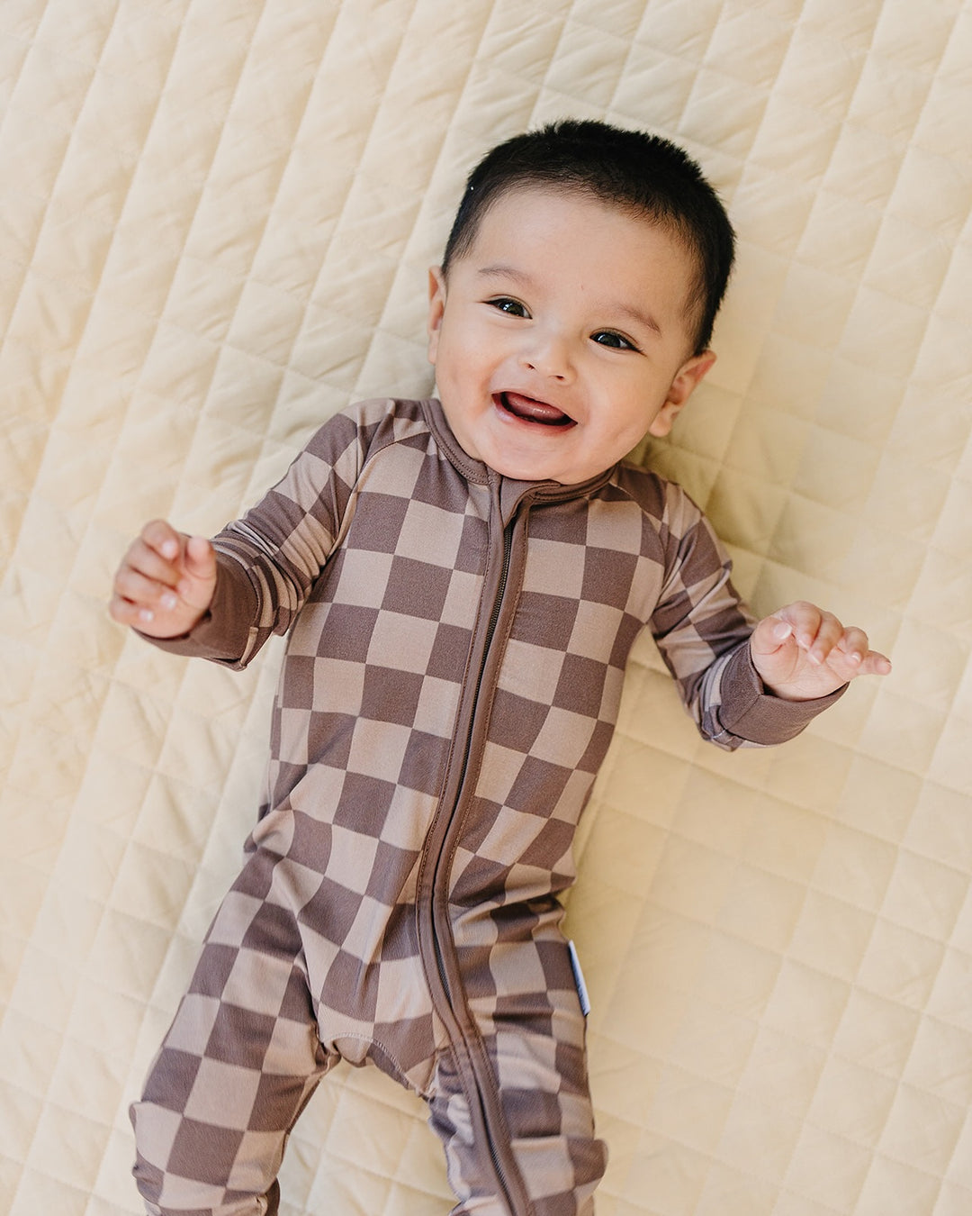 A smiling baby lying on a quilted cream blanket, wearing a brown and beige checkered zip-up footless one-piece pajama.