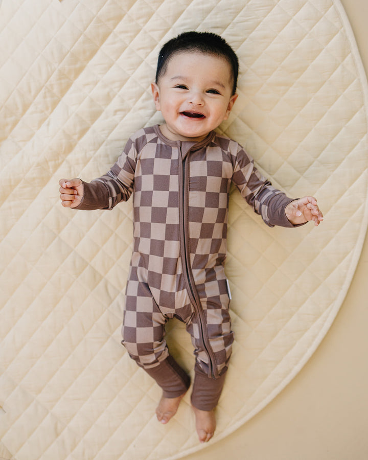 Smiling baby lying on a cream quilt in the Espresso Checkered romper with arms open and feet uncovered.