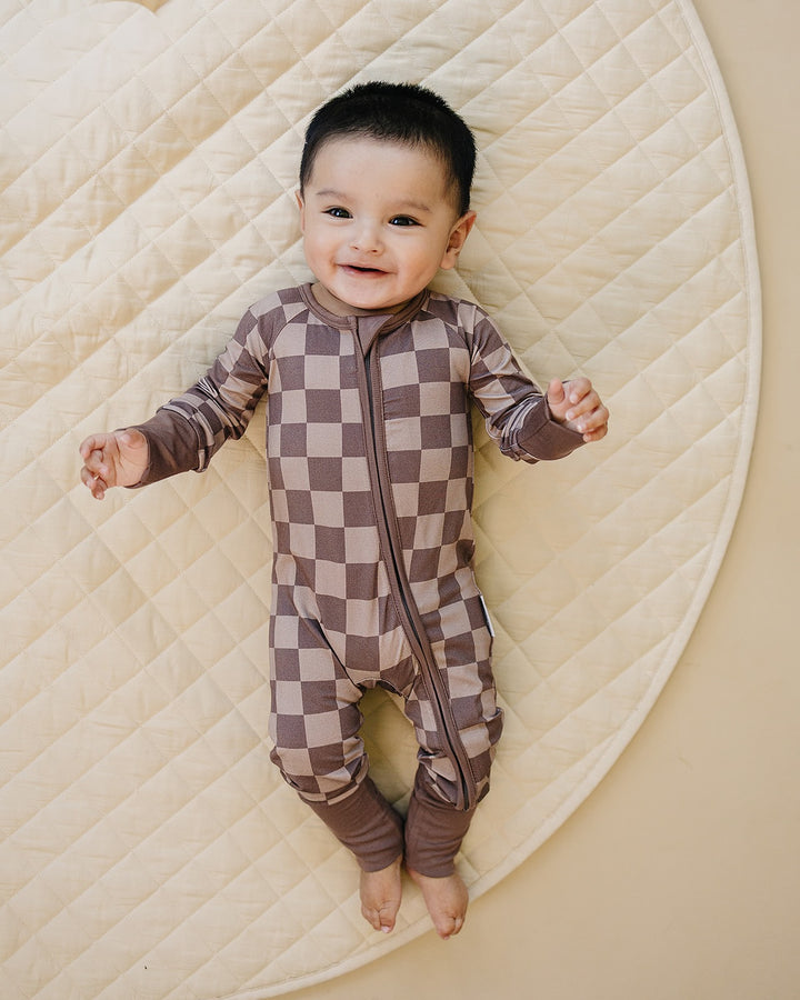 A baby lying on a cream quilted blanket, wearing a brown and beige checkered one-piece footed sleeper with a front zipper.