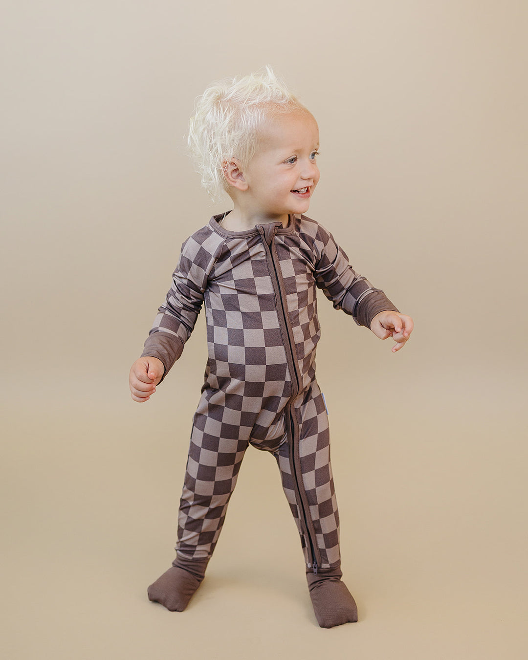 A toddler wearing a long-sleeve, one-piece footed pajama with a brown and gray checkerboard pattern, standing against a solid beige studio background.