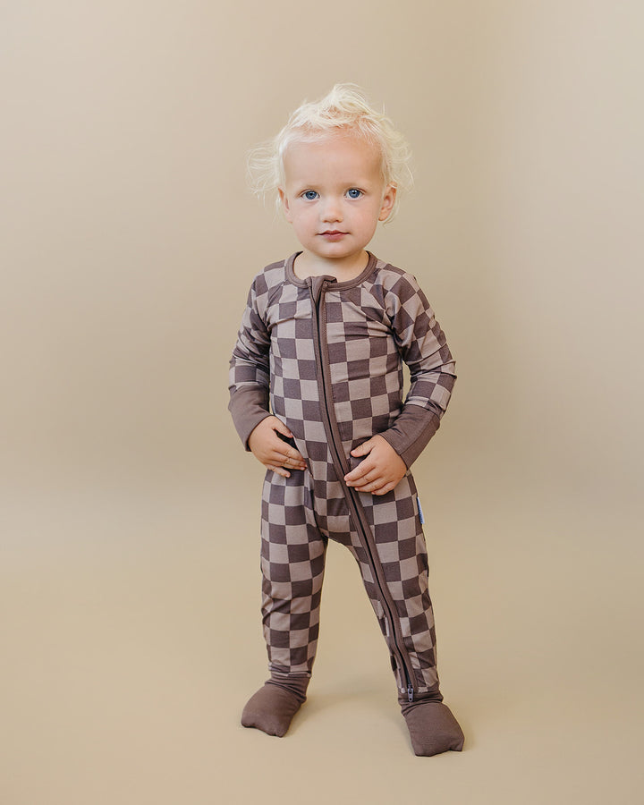 A toddler with blonde hair stands wearing a brown and gray checkerboard zip-up footed one-piece pajama against a solid beige studio background.