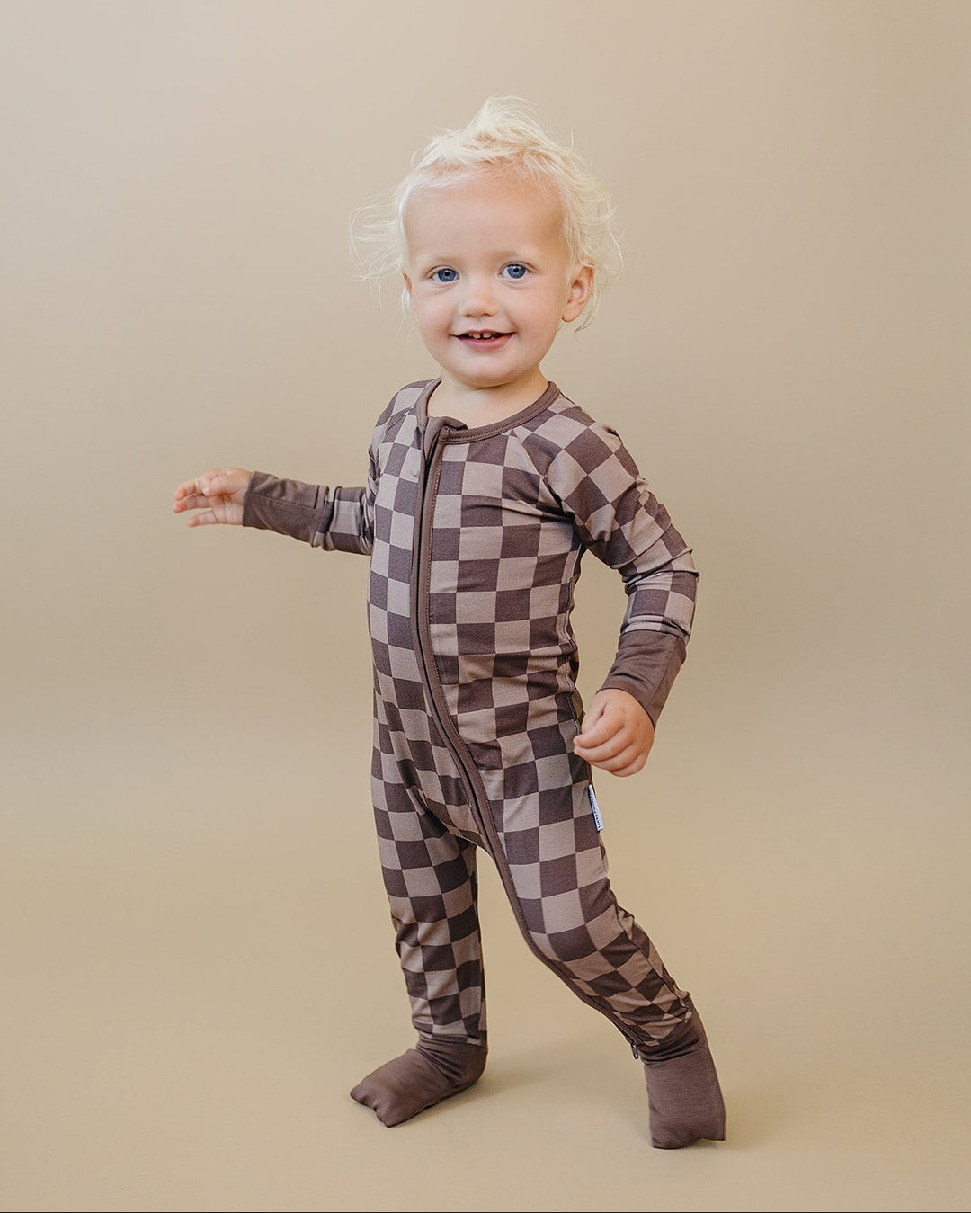 Smiling toddler standing in the Espresso Checkered zip romper with brown cuffs and covered feet.