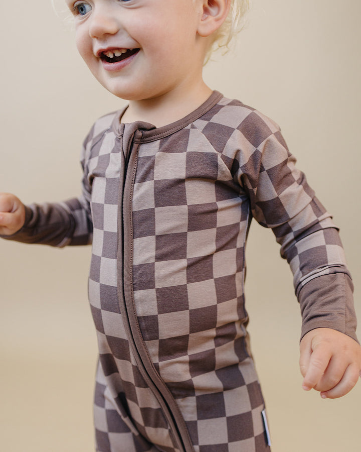 A smiling toddler wearing a long-sleeve one-piece pajama with a brown and beige checkerboard pattern, standing against a plain beige background.