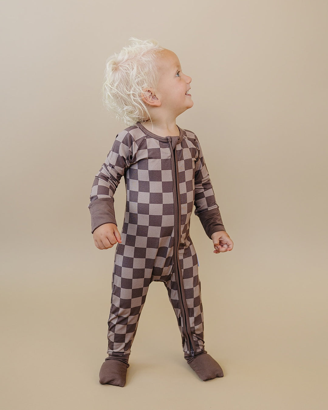 Toddler looking up while wearing the Espresso Checkered zip romper with brown and taupe squares and covered feet.