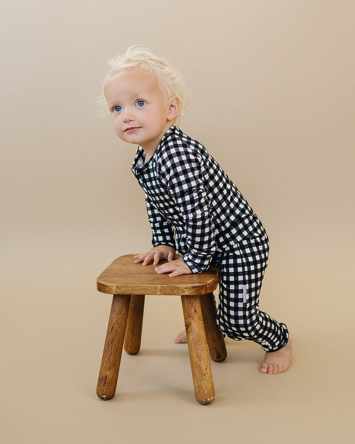 Little one wearing the Black Gingham Bamboo Zip Romper beside a wooden stool