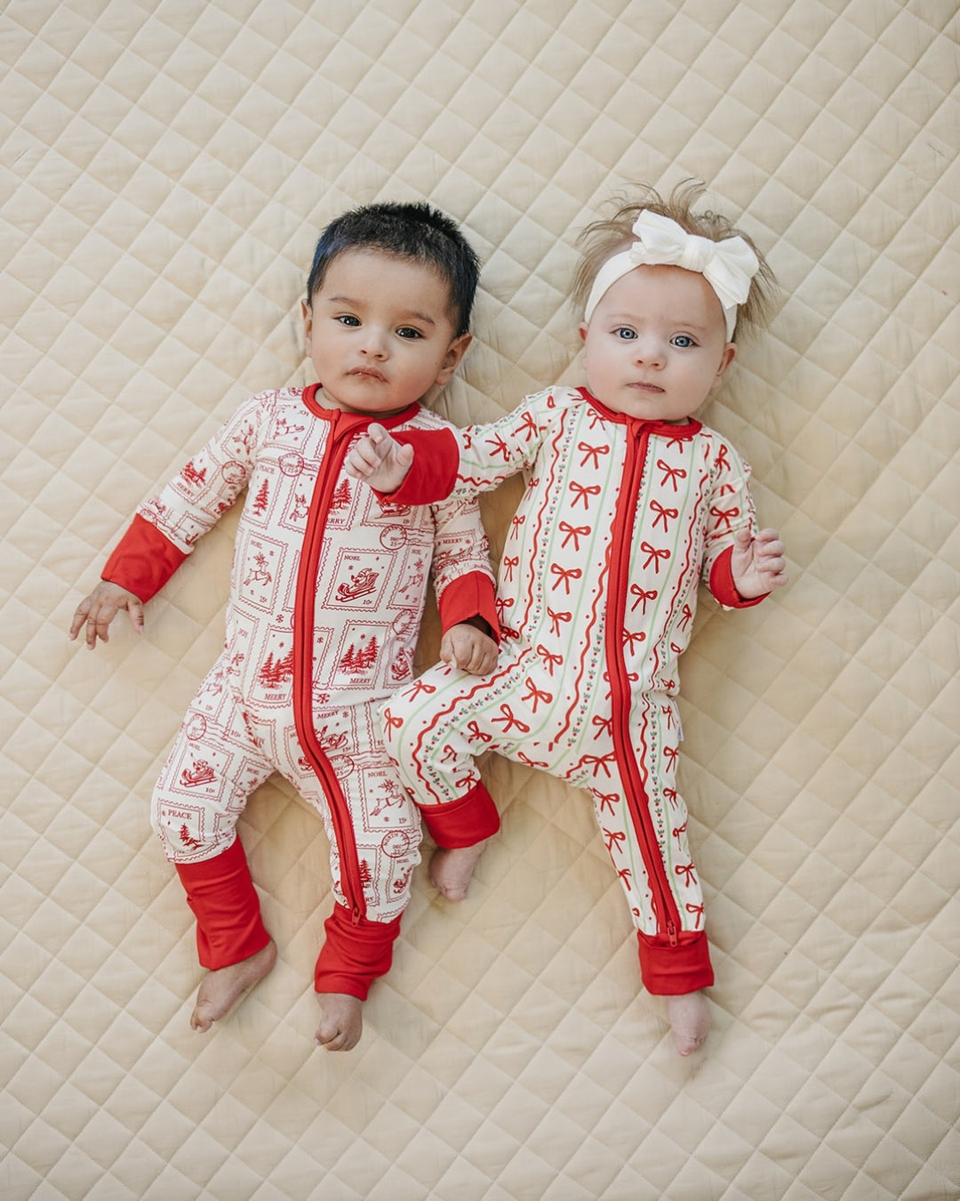 Two babies lying beside each other on a quilt in matching North Pole Stamps Christmas zip rompers.