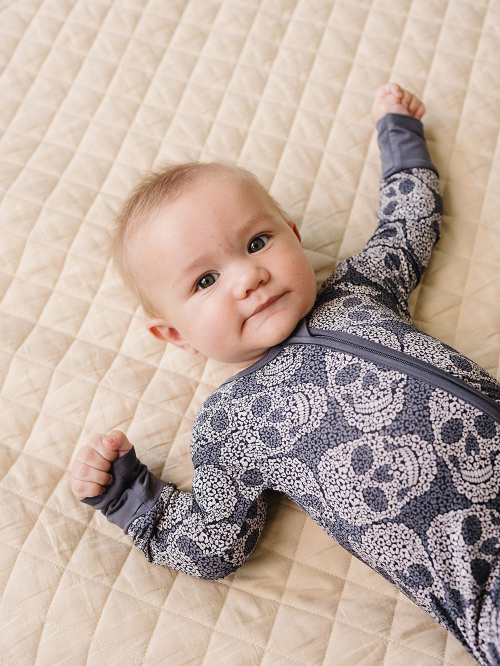 Smiling baby wearing the Leopard Skull Bamboo Zip Romper on a quilted blanket.