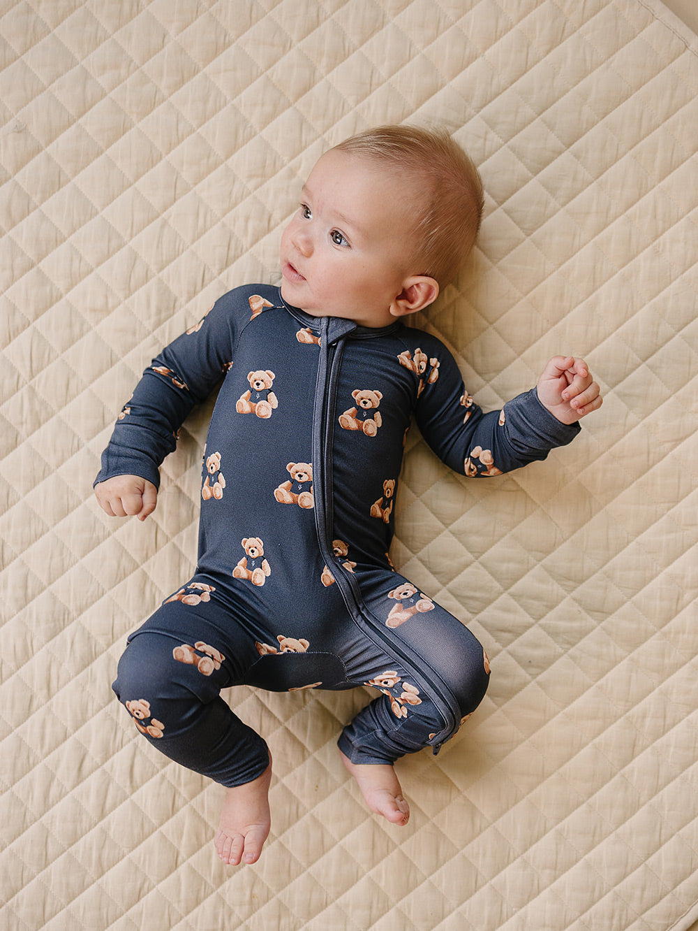 Baby lying on a cream quilted mat in the dark charcoal teddy bear-print romper.