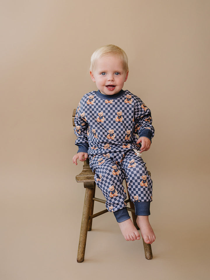 Smiling baby modeling the blue checkered bear-print jogger set on a wooden stool.