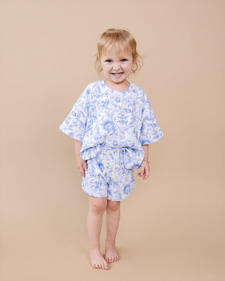 A smiling toddler stands barefoot against a solid beige studio background, wearing a light blue and white floral print two-piece outfit consisting of a short-sleeve tunic top and matching shorts.