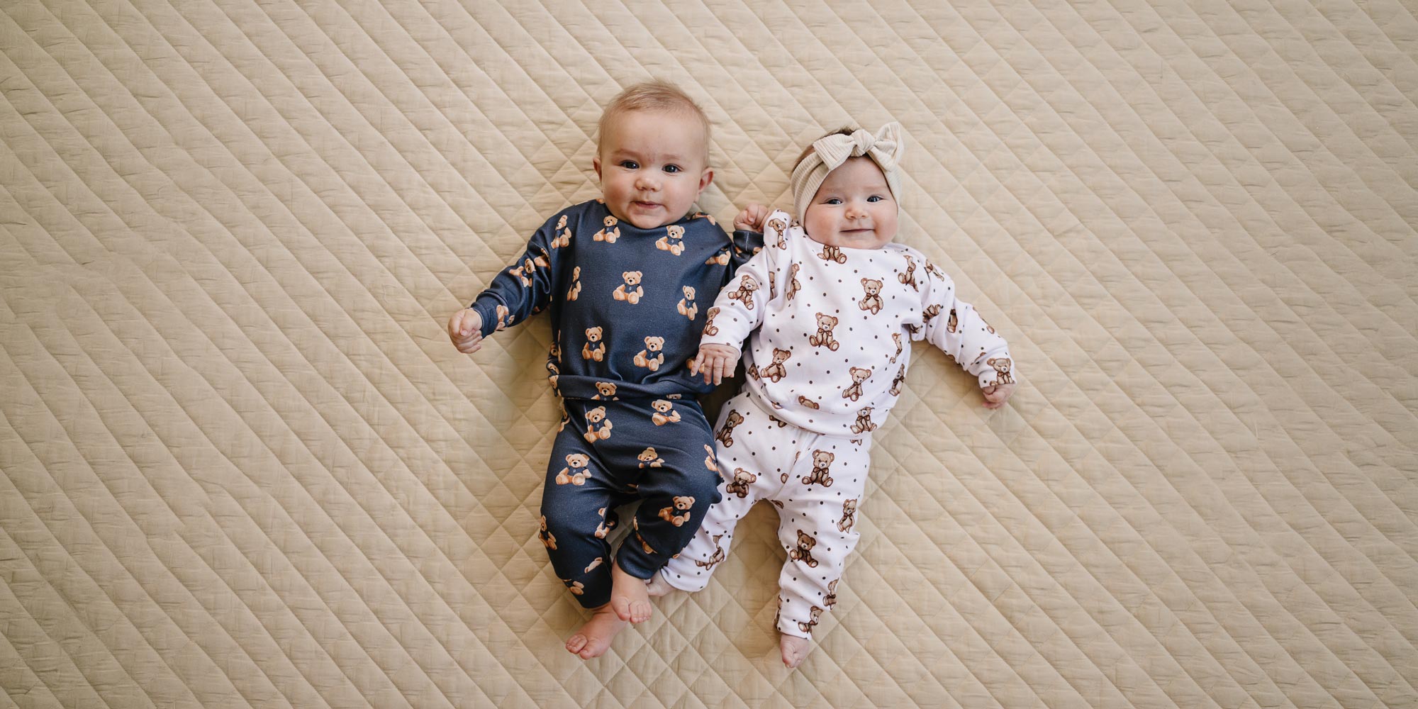 Two babies lying on a cream quilt wearing coordinating dark charcoal and white teddy bear-print outfits.