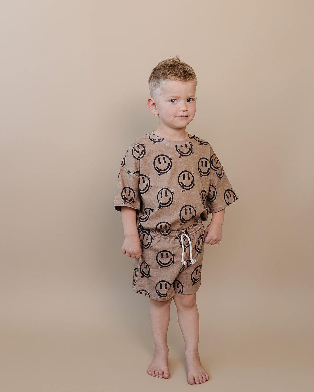 toddler boy wearing Lucky Panda Kids Smiley Brown Shorts Set — warm brown all-over smiley face print cotton two-piece outfit