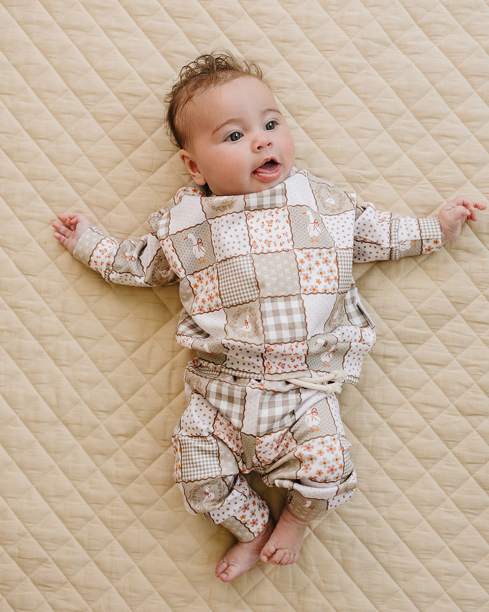 Baby girl lying with her arms out in the Cottage Goose two-piece jogger set on a soft beige quilt.