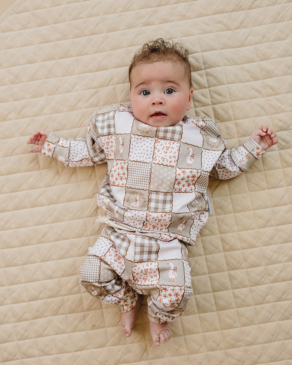 Baby girl lying on a beige quilt wearing the Lucky Panda Kids Cottage Goose cotton jogger set in a neutral patchwork print.