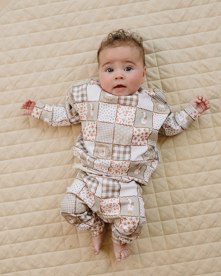 Baby girl lying on a beige quilt wearing the Lucky Panda Kids Cottage Goose cotton jogger set in a neutral patchwork print.