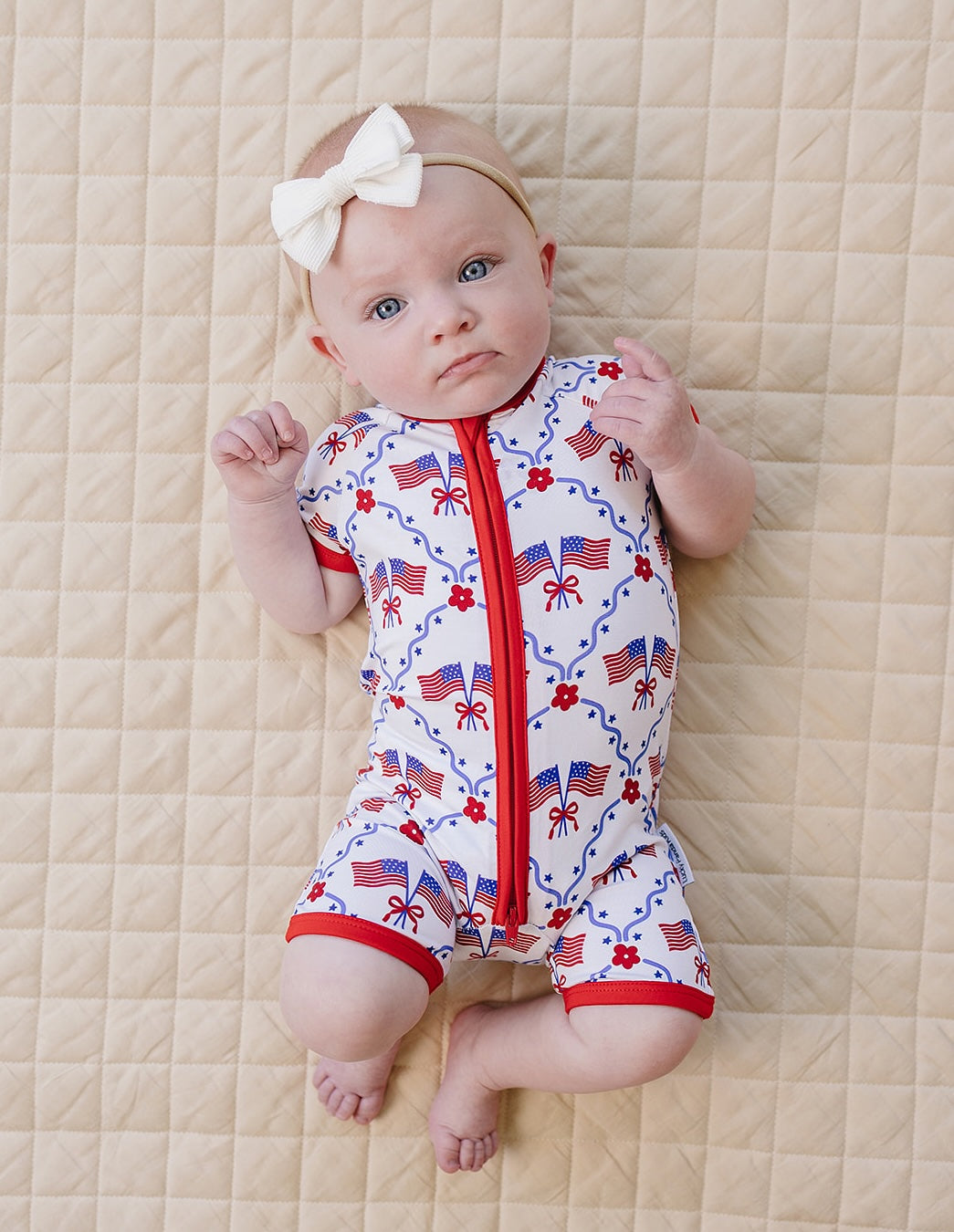 Baby wearing a red, white, and blue onesie with American flag pattern on a beige quilted surface.
