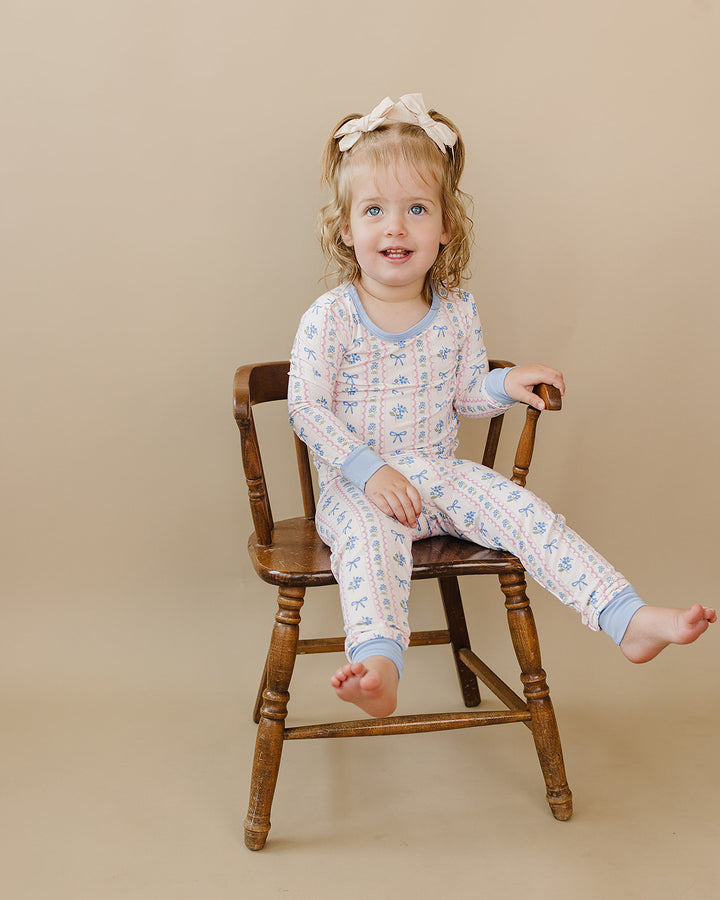 Toddler seated on a wooden chair with one leg extended in the Whimsy Bloom floral bamboo two-piece set.