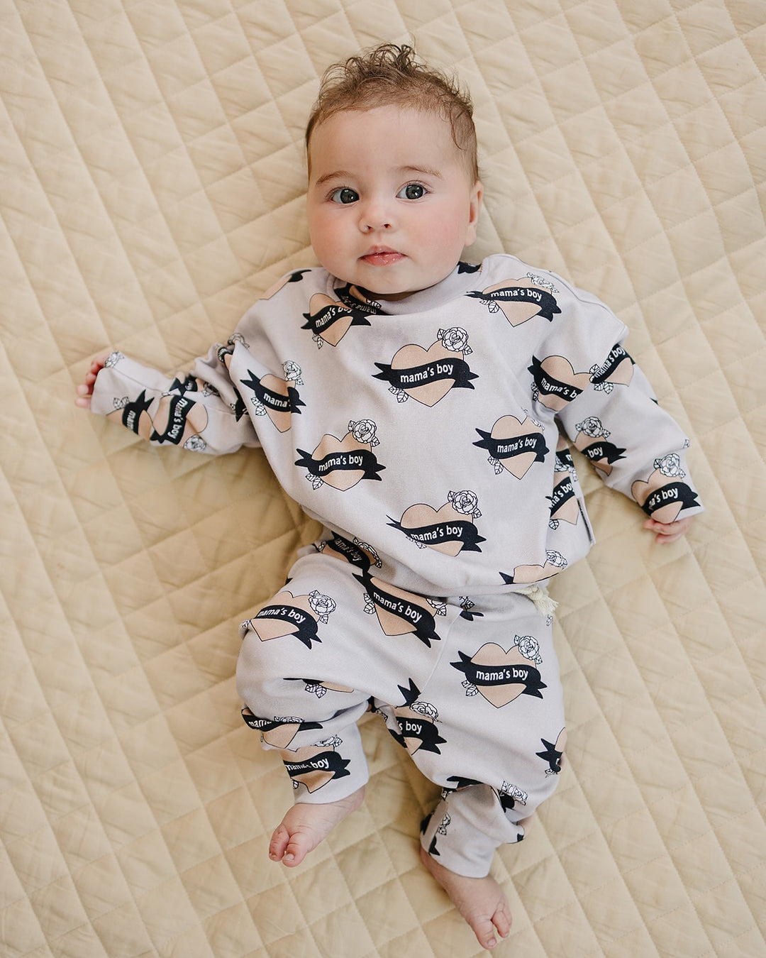 Baby lying on a cream quilt in the Mama’s Boy cotton jogger set with beige hearts and black banners.