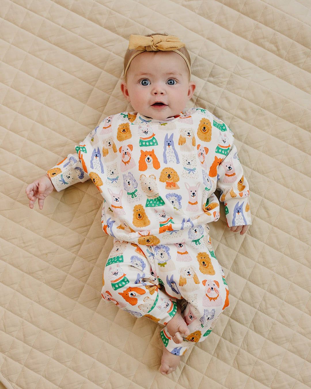 Smiling baby wearing the Whimsy Bloom bamboo zip romper and white bow headband on a cream quilt.