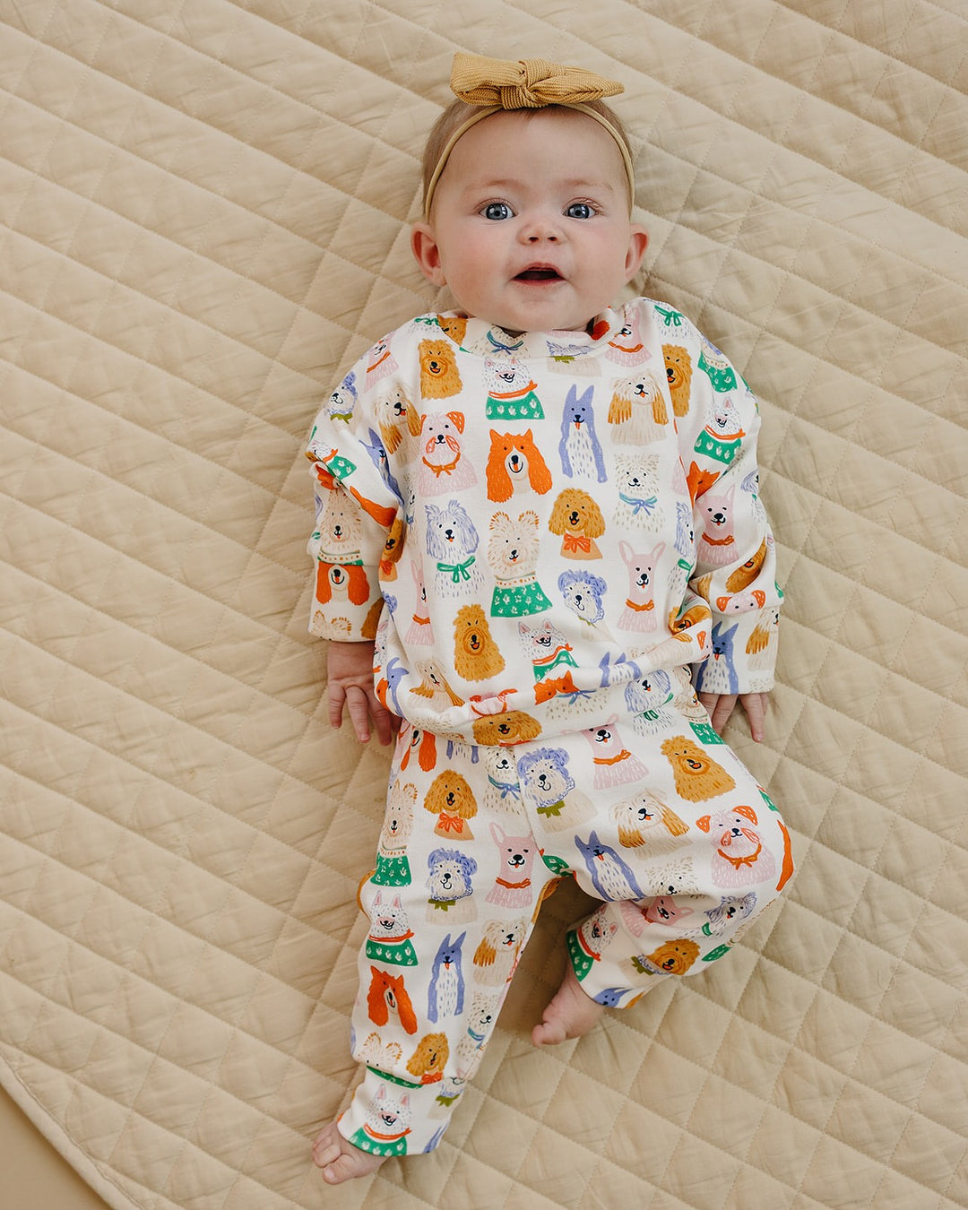 Baby lying on a cream quilt in the Puppy Club cotton jogger set with colorful dog illustrations and a mustard bow headband.