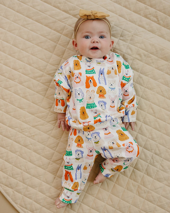 Baby lying on a cream quilt in the Puppy Club cotton jogger set with colorful dog illustrations and a mustard bow headband.