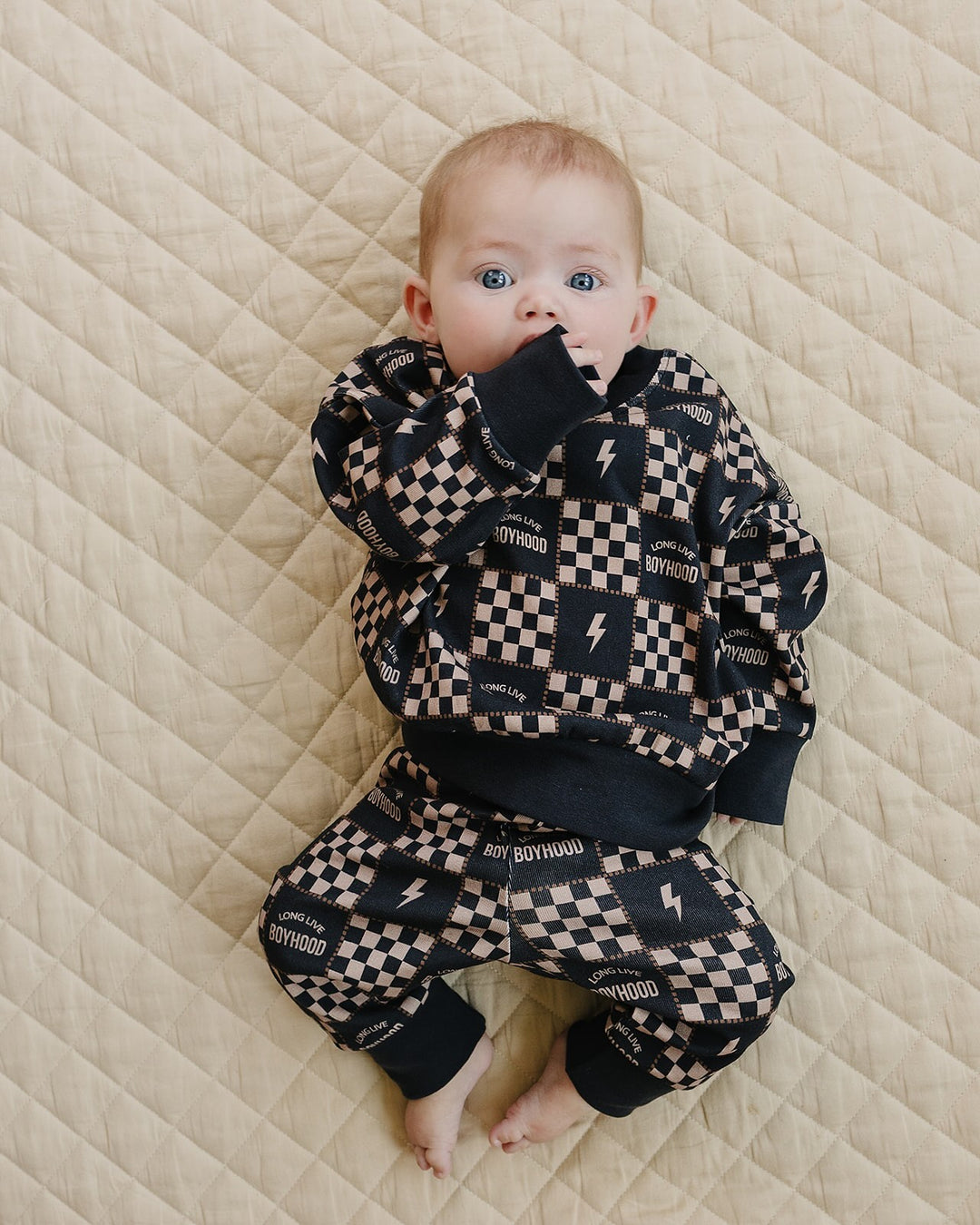 Baby lying on a cream quilt in the Long Live Boyhood cotton jogger set with charcoal and beige checks and lightning bolts.