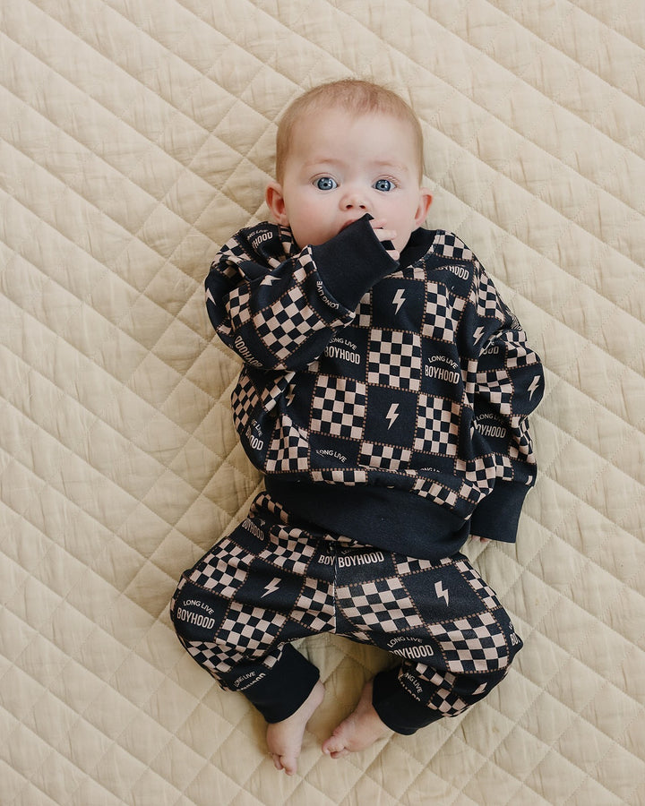 Baby lying on a cream quilt in the Long Live Boyhood cotton jogger set with charcoal and beige checks and lightning bolts.
