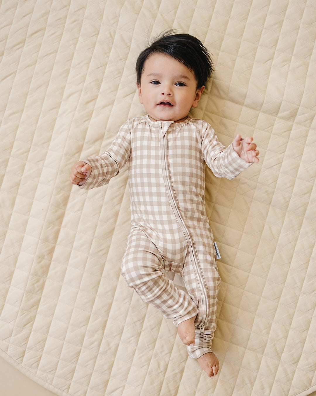 Full-body view of a baby wearing the Beige Gingham bamboo zip romper on a quilted blanket.