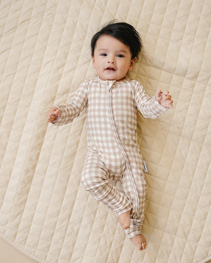 Full-body view of a baby wearing the Beige Gingham bamboo zip romper on a quilted blanket.