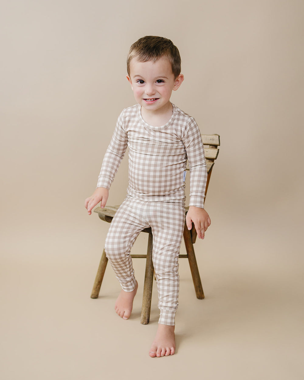 Toddler wearing the Beige Gingham bamboo two-piece set beside a wooden stool.