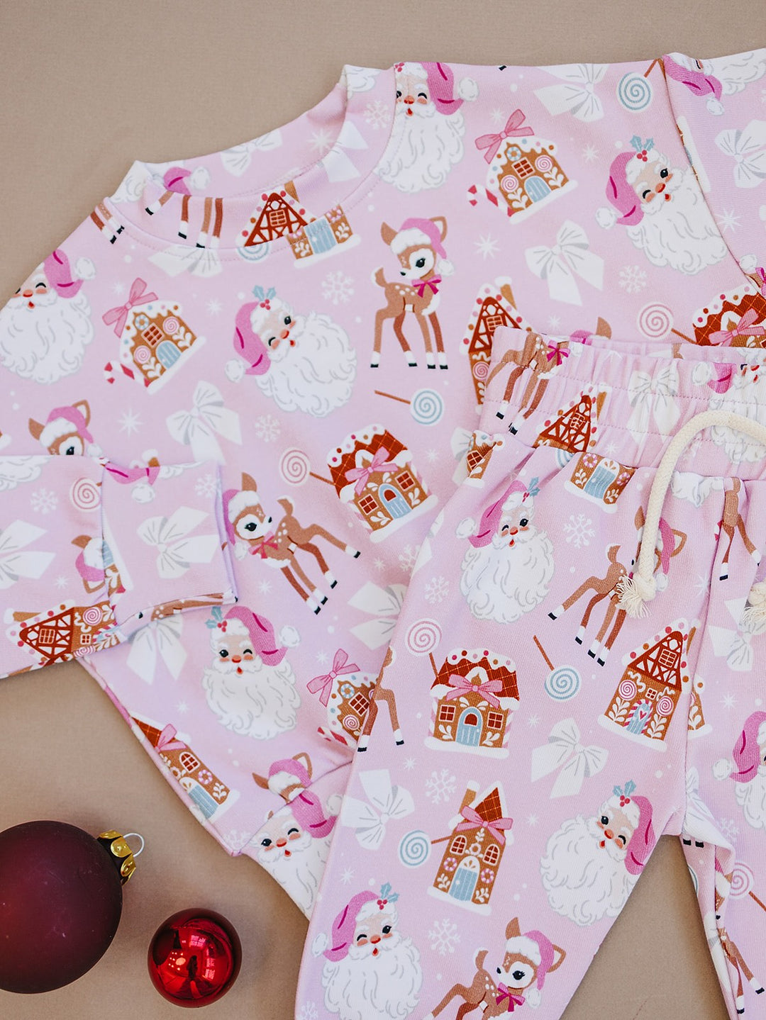 Detail of the Pink Christmas jogger set showing Santa, reindeer and gingerbread houses on pink fabric beside
ornaments.